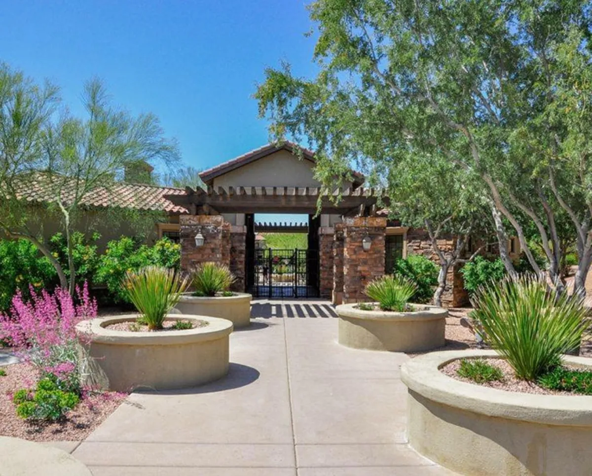 A welcoming gated entrance framed by lush landscaped gardens and colorful flower beds. A winding pathway leads toward the entrance, conveying a sense of privacy and exclusivity within a safe, serene environment.