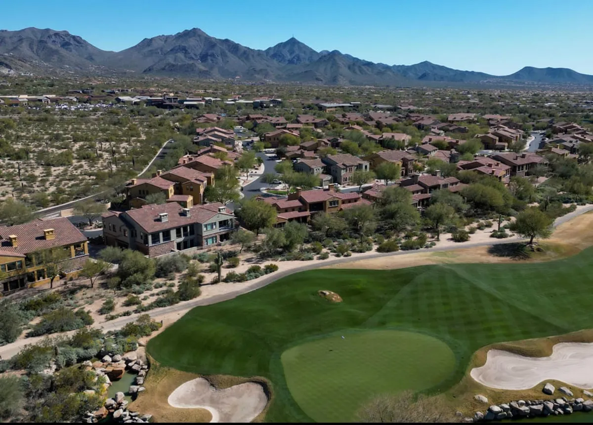 A panoramic view of a lush golf course with manicured fairways set against a dramatic mountain backdrop. The image also captures a glimpse of nearby residential areas, highlighting the seamless integration of luxury living and outdoor recreation.