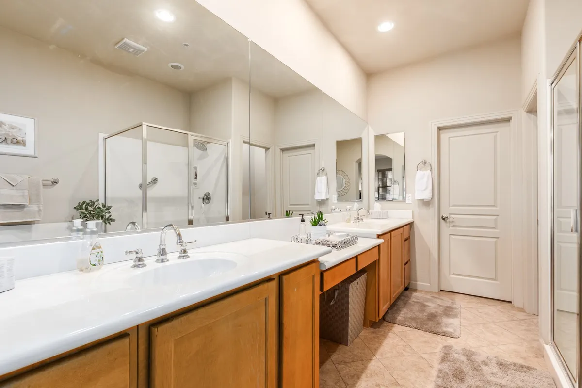 A modern bathroom with dual sinks, large mirrors, a spacious shower stall, elegant wood cabinetry, and a decorative plant. The setting is orderly and serene, perfect for morning routines or unwinding after the day.